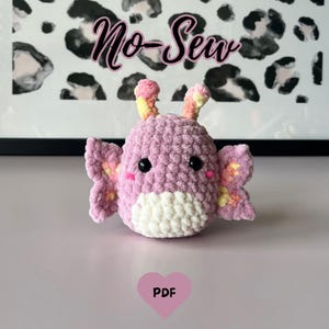 Op de afbeelding: Een handgemaakt, pluche, roze en wit vlindervormig speelgoed met zwarte ogen en roze wangen. Het speelgoed heeft gele accenten op de vleugels en antennes. De woorden "No-Sew" staan boven het speelgoed.