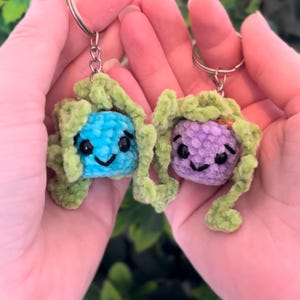 May include: Two handmade crochet keychains, one blue and one purple, each with a smiling face and green accents. The keychains are held in open hands against a blurred green background, showcasing a whimsical design.
