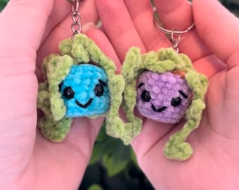 May include: Two handmade crochet keychains, one blue and one purple, each with a smiling face and green accents. The keychains are held in open hands against a blurred green background, showcasing a whimsical design.