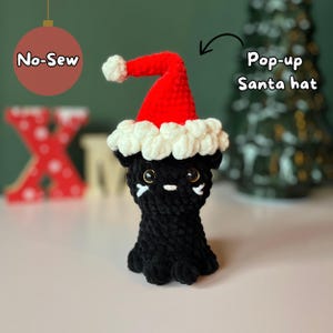 Puede incluir: Gato negro de crochet con un gorro de Papá Noel emergente rojo y blanco. El gorro tiene un pompón blanco y las palabras "No-Sew" son visibles en el fondo.