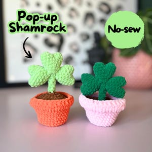 May include: Two crocheted shamrock plants in pots. The shamrock on the left is green and in an orange pot. The shamrock on the right is green and in a pink pot. The text "Pop-up Shamrock" is in a green circle above the left shamrock. The text "No-sew" is in a green circle above the right shamrock.
