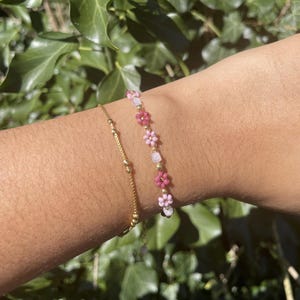May include: Two bracelets on a wrist. One is a delicate gold chain with small beads. The other is a beaded bracelet with pink and white flower-shaped beads. The background is green foliage.