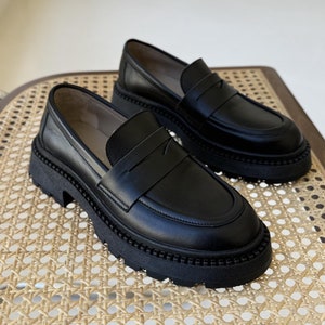 May include: A pair of black leather loafers with a chunky platform sole. The loafers have a simple design with a single strap across the vamp.