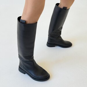 May include: A pair of black leather knee-high boots with a low heel. The boots have a simple design and are made of a smooth leather.