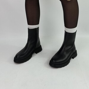 Könnte beinhalten: Schwarze Leder-Stiefeletten mit einer dicken Sohle und einem Reißverschluss an der Seite. Die Stiefeletten werden mit schwarzen Strumpfhosen und einem braunen Mantel getragen.