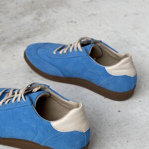 May include: A pair of blue suede trainers with white leather accents and brown rubber soles. The trainers are on a light grey surface.