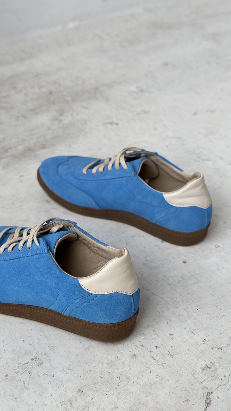 May include: A pair of blue suede trainers with white leather accents and brown rubber soles. The trainers are on a light grey surface.