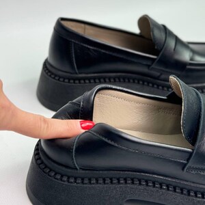 Puede incluir: Un par de mocasines de cuero negro con una suela de plataforma gruesa. Los mocasines tienen un dise&ntilde;o cl&aacute;sico de mocas&iacute;n penny con una tira en la parte superior.