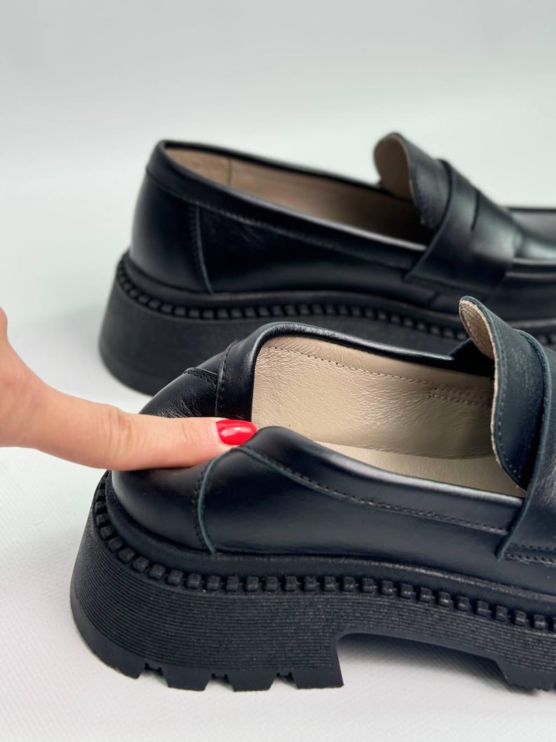 Puede incluir: Un par de mocasines de cuero negro con una suela de plataforma gruesa. Los mocasines tienen un dise&ntilde;o cl&aacute;sico de mocas&iacute;n penny con una tira en la parte superior.