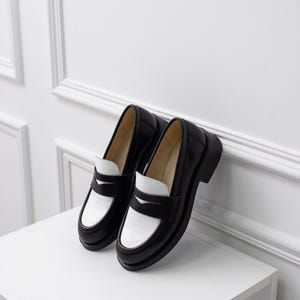 K&ouml;nnte beinhalten: Ein Paar schwarze und wei&szlig;e Leder-Slipper mit kontrastierendem Farbdesign. Die Slipper haben eine abgerundete Spitze und einen dicken Absatz.