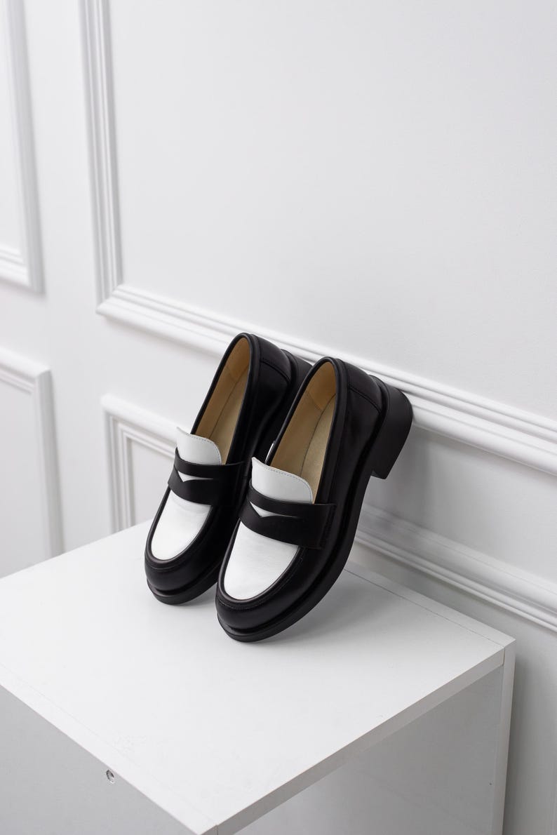 K&ouml;nnte beinhalten: Ein Paar schwarze und wei&szlig;e Leder-Slipper mit kontrastierendem Farbdesign. Die Slipper haben eine abgerundete Spitze und einen dicken Absatz.