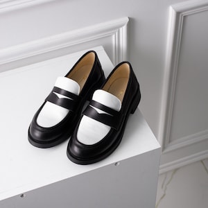 K&ouml;nnte beinhalten: Zwei schwarze Leder-Slipper mit wei&szlig;en Ledereins&auml;tzen. Die Slipper haben eine abgerundete Spitze und einen niedrigen Absatz.
