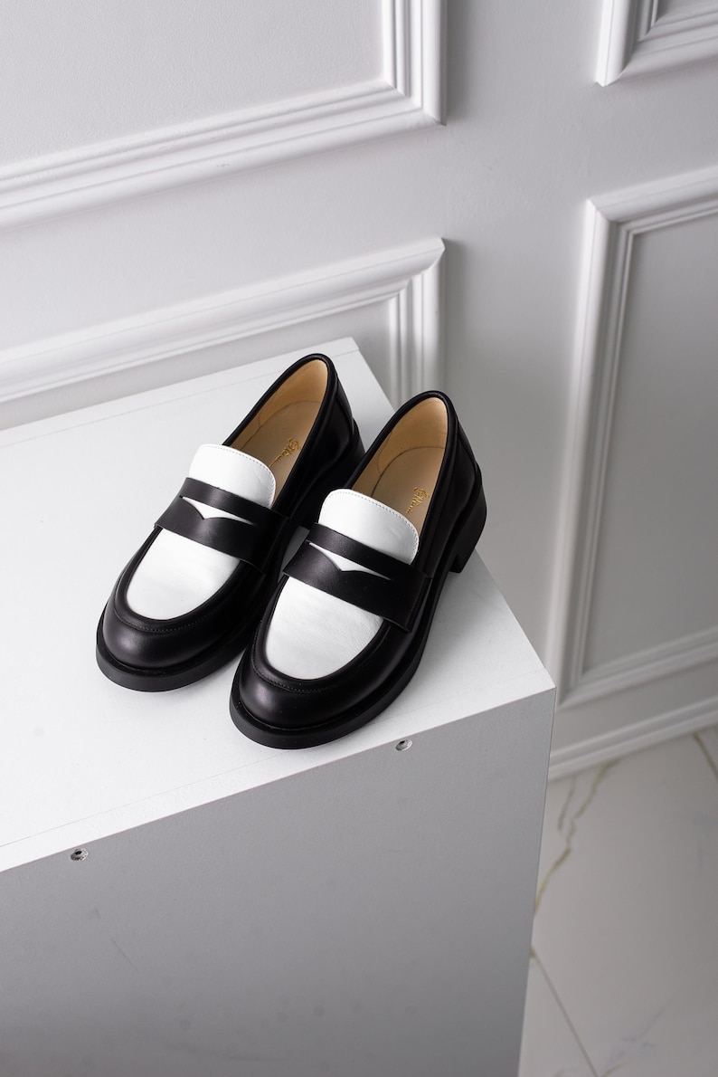 K&ouml;nnte beinhalten: Zwei schwarze Leder-Slipper mit wei&szlig;en Ledereins&auml;tzen. Die Slipper haben eine abgerundete Spitze und einen niedrigen Absatz.