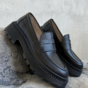 Puede incluir: Un par de mocasines de cuero negro con suela de plataforma gruesa. Los mocasines tienen un dise&ntilde;o cl&aacute;sico de mocas&iacute;n penny con una tira en la parte superior.