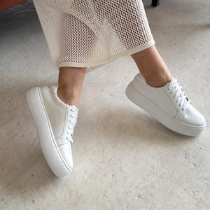 Può includere: Sneakers bianche in pelle con piattaforma e lacci. Le sneakers hanno una suola bianca spessa e sono indossate da una persona che indossa una gonna in rete bianca.