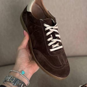 May include: Brown suede sneakers with white laces and a cream coloured trim around the top of the shoe.