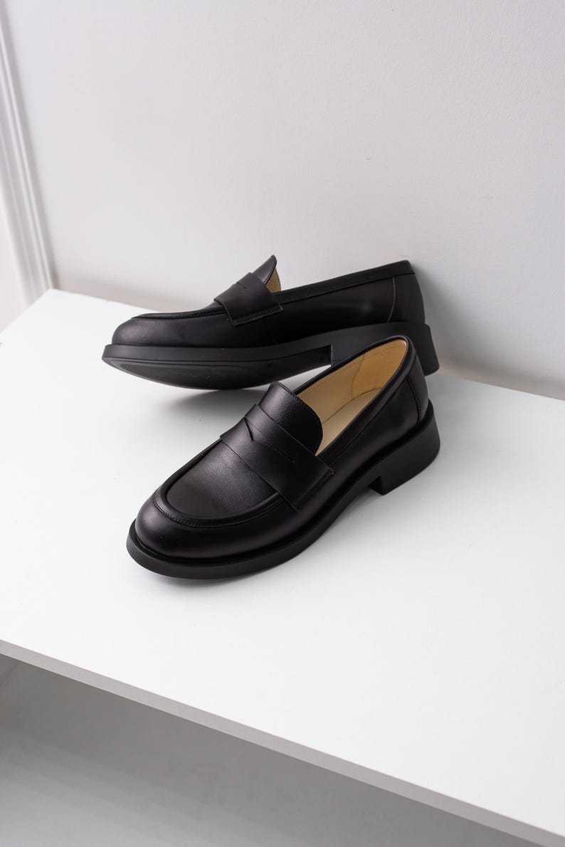 K&ouml;nnte beinhalten: Ein Paar schwarze Leder-Slipper mit klassischem Penny-Riemen-Design. Die Slipper haben eine leicht klobige Sohle und eine abgerundete Spitze.