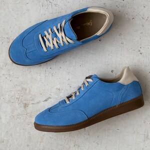 May include: A pair of blue suede trainers with white laces and a brown rubber sole. The trainers have a cream-coloured lining.