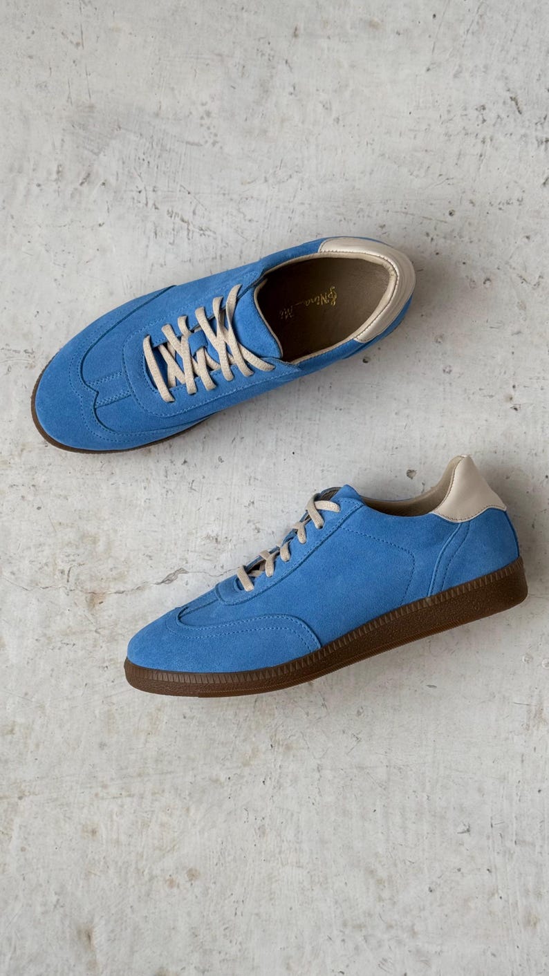 May include: A pair of blue suede trainers with white laces and a brown rubber sole. The trainers have a cream-coloured lining.