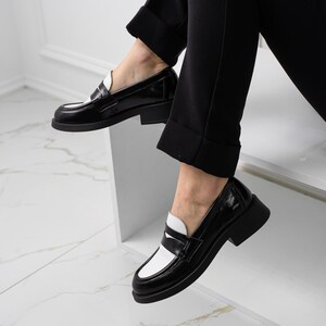 K&ouml;nnte beinhalten: Schwarz-wei&szlig;e Leder-Slipper mit kontrastierendem Farbdesign. Die Slipper haben eine abgerundete Spitze und einen niedrigen Blockabsatz.