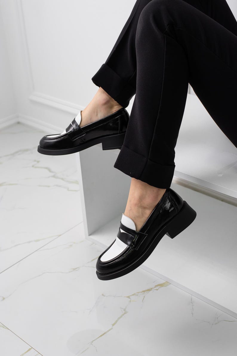 K&ouml;nnte beinhalten: Schwarz-wei&szlig;e Leder-Slipper mit kontrastierendem Farbdesign. Die Slipper haben eine abgerundete Spitze und einen niedrigen Blockabsatz.