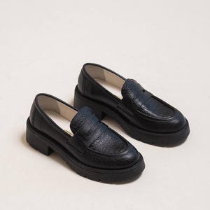 May include: A pair of black leather loafers with a textured, crocodile-like pattern. The shoes have a chunky sole and a classic penny loafer design. The image shows the shoes from a slightly elevated angle, showcasing their details.