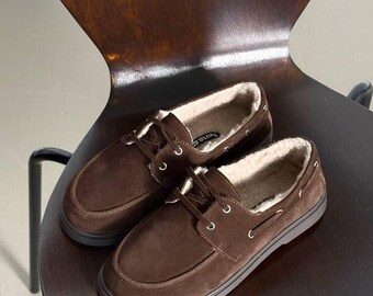 Op de afbeelding: Een paar bruine suède bootschoenen met een imitatiebontvoering. De schoenen hebben een vetersluiting en een rubberen zool. De schoenen worden gepresenteerd op een houten stoel.
