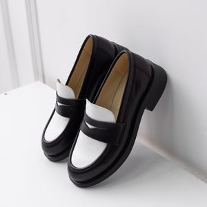 K&ouml;nnte beinhalten: Ein Paar schwarze und wei&szlig;e Leder-Slipper mit kontrastierendem Farbdesign. Die Slipper haben eine klobige Plateau-Sohle und einen schlichten, klassischen Stil.