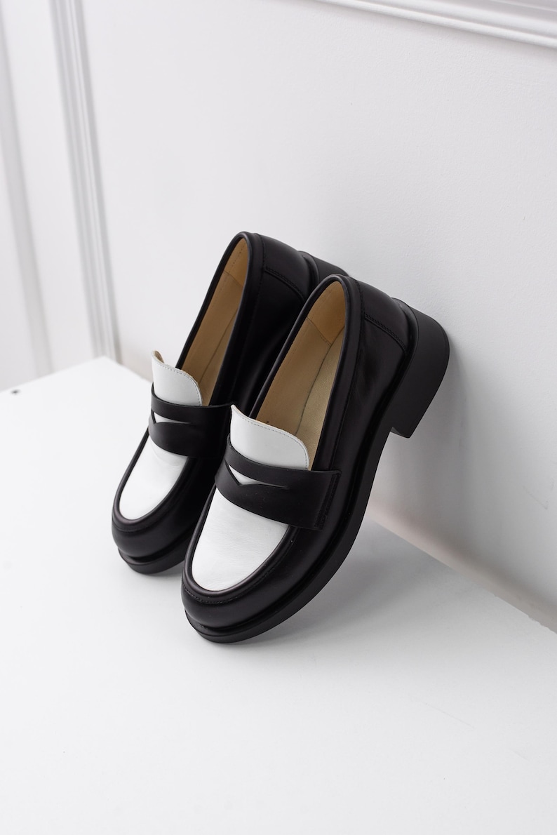 K&ouml;nnte beinhalten: Ein Paar schwarze und wei&szlig;e Leder-Slipper mit kontrastierendem Farbdesign. Die Slipper haben eine klobige Plateau-Sohle und einen schlichten, klassischen Stil.