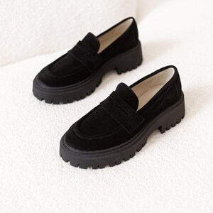 May include: Black suede loafers with a chunky platform sole. The loafers feature a classic penny loafer design with a strap across the top.