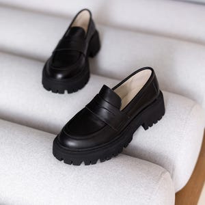 Puede incluir: Un par de mocasines de cuero negro con suela de plataforma gruesa. Los mocasines tienen un dise&ntilde;o cl&aacute;sico de mocas&iacute;n penny con una tira en la parte superior.