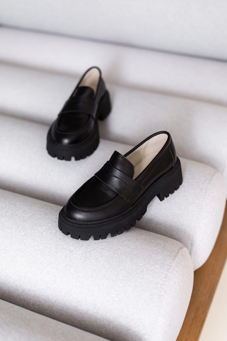 Puede incluir: Un par de mocasines de cuero negro con suela de plataforma gruesa. Los mocasines tienen un dise&ntilde;o cl&aacute;sico de mocas&iacute;n penny con una tira en la parte superior.