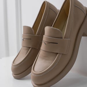 K&ouml;nnte beinhalten: Ein Paar hellbraune Lederschuhe mit einem schlichten Design. Die Schuhe haben eine abgerundete Spitze und eine Naht um den Rand der Sohle.