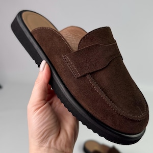 May include: Brown suede leather slip-on loafers with a black rubber sole. The loafers have a simple design with a single strap across the top.