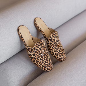 May include: A pair of leopard print slip-on mules with a gold buckle detail.