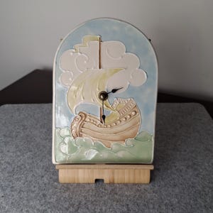 May include: An arched ceramic clock featuring a relief design of a sailing ship on a light blue sky and green sea background. The ship has cream-coloured sails and a tan hull. Black clock hands are centred on the ship's sail and mast.