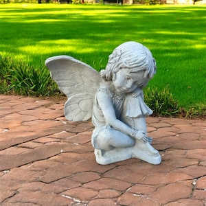May include: A gray concrete fairy statue with large, detailed wings. The fairy is depicted in a seated position, with its head bowed and hands clasped. The statue is on a brick patio with a green lawn in the background.