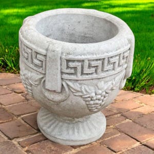 May include: A gray concrete planter with a wide, open top. The planter features a Greek key design around the rim and a grape and leaf motif. It has a pedestal base and two handles. The planter is outdoors on a brick surface.