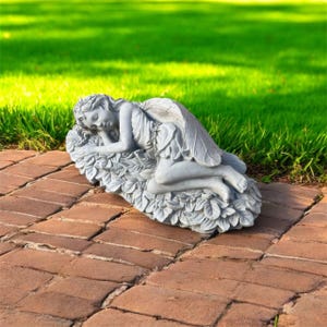 May include: A gray stone fairy statue resting on a bed of leaves. The fairy is depicted with closed eyes and delicate wings, wearing a flowing dress. The statue is placed on a brick surface with a green grassy background.