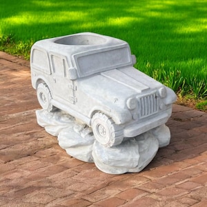 May include: A gray concrete planter shaped like a vintage jeep. The planter has a rectangular opening for plants and sits on a rock-like base. The jeep features detailed tires, a front grill, and a roof.
