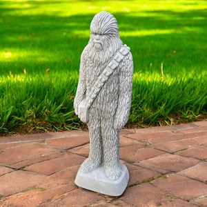 May include: A gray concrete statue of a Wookiee character, standing on a rectangular base. The statue has a detailed fur texture, a bandolier, and a stern expression. The statue is outdoors on a brick patio with green grass in the background.