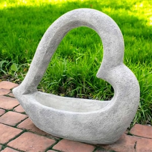 May include: A gray, heart-shaped planter made of stone. The planter has a textured surface and a wide opening for plants. The heart design is open in the center, creating a unique and decorative garden accent.