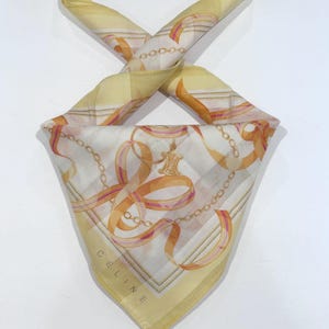 May include: A square silk scarf with a repeating pattern of gold chains and ribbons on a white background. The scarf has a yellow border and the word "CELINE" is printed in the corner.