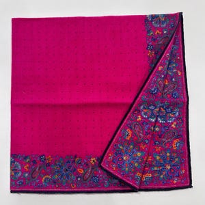 Puede incluir: Un pa&ntilde;uelo de algod&oacute;n rosa con un borde de patr&oacute;n paisley en tonos de azul, verde y amarillo. El pa&ntilde;uelo est&aacute; doblado por la mitad y tiene un ribete azul oscuro.