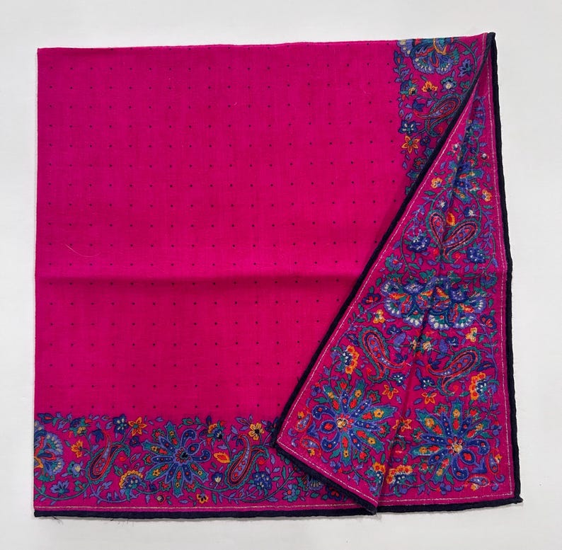Puede incluir: Un pa&ntilde;uelo de algod&oacute;n rosa con un borde de patr&oacute;n paisley en tonos de azul, verde y amarillo. El pa&ntilde;uelo est&aacute; doblado por la mitad y tiene un ribete azul oscuro.