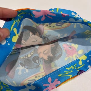 May include: A blue fabric item with a colorful pattern featuring cartoon characters. The design includes images of a blue alien-like creature and a young girl, set against a backdrop of splashes and shapes.
