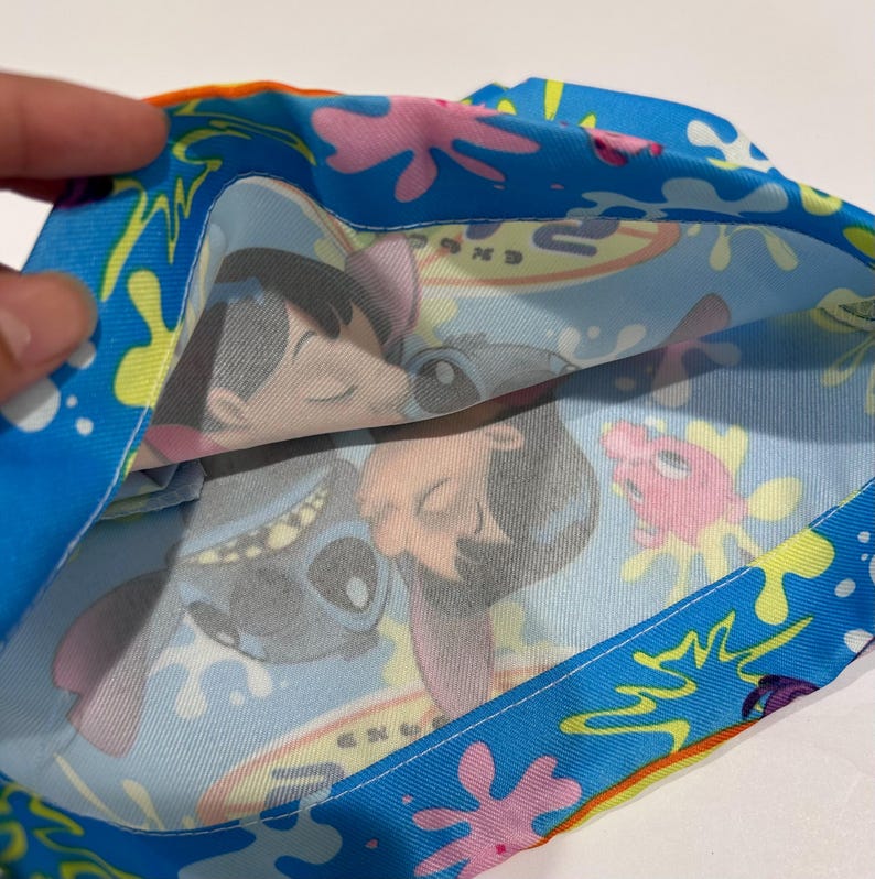 May include: A blue fabric item with a colorful pattern featuring cartoon characters. The design includes images of a blue alien-like creature and a young girl, set against a backdrop of splashes and shapes.