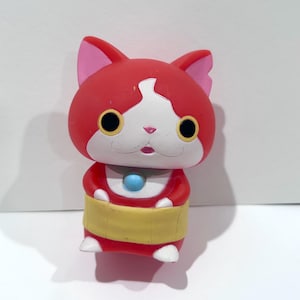 May include: A red and white cartoon cat figurine with large yellow eyes and pink ears. The cat has a yellow band around its waist and a blue gem on its chest. The figurine is standing upright.