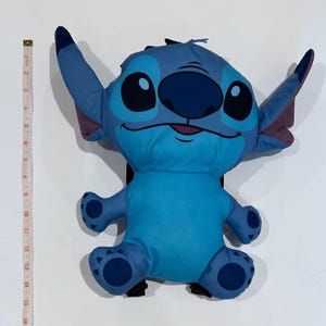 May include: A blue Stitch backpack with large ears, black eyes, and a dark blue nose. The backpack is approximately 16 inches tall. The design is based on the character from the Disney movie Lilo & Stitch. The backpack has a black back and straps.