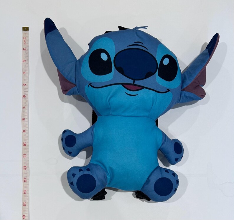 May include: A blue Stitch backpack with large ears, black eyes, and a dark blue nose. The backpack is approximately 16 inches tall. The design is based on the character from the Disney movie Lilo & Stitch. The backpack has a black back and straps.